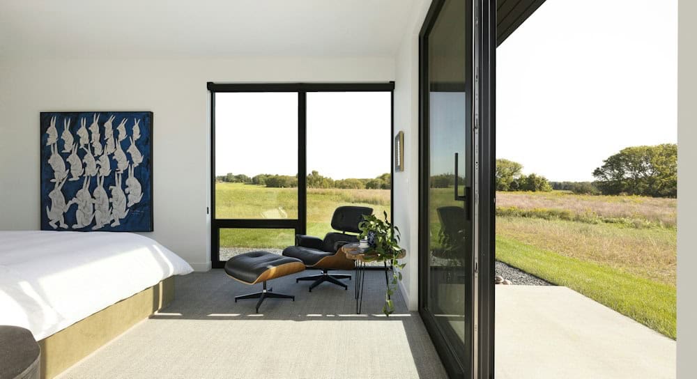 Primary bedroom with a white bed, blue rabbit artwork and an Eames lounge chair by windows, with a sliding door open to the prairie