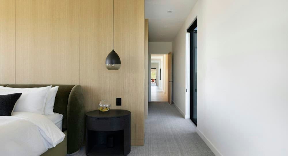 Olive velvet bed against a white oak paneled wall with a black glass pendant over a round black nightstand, beside a carpeted hallway