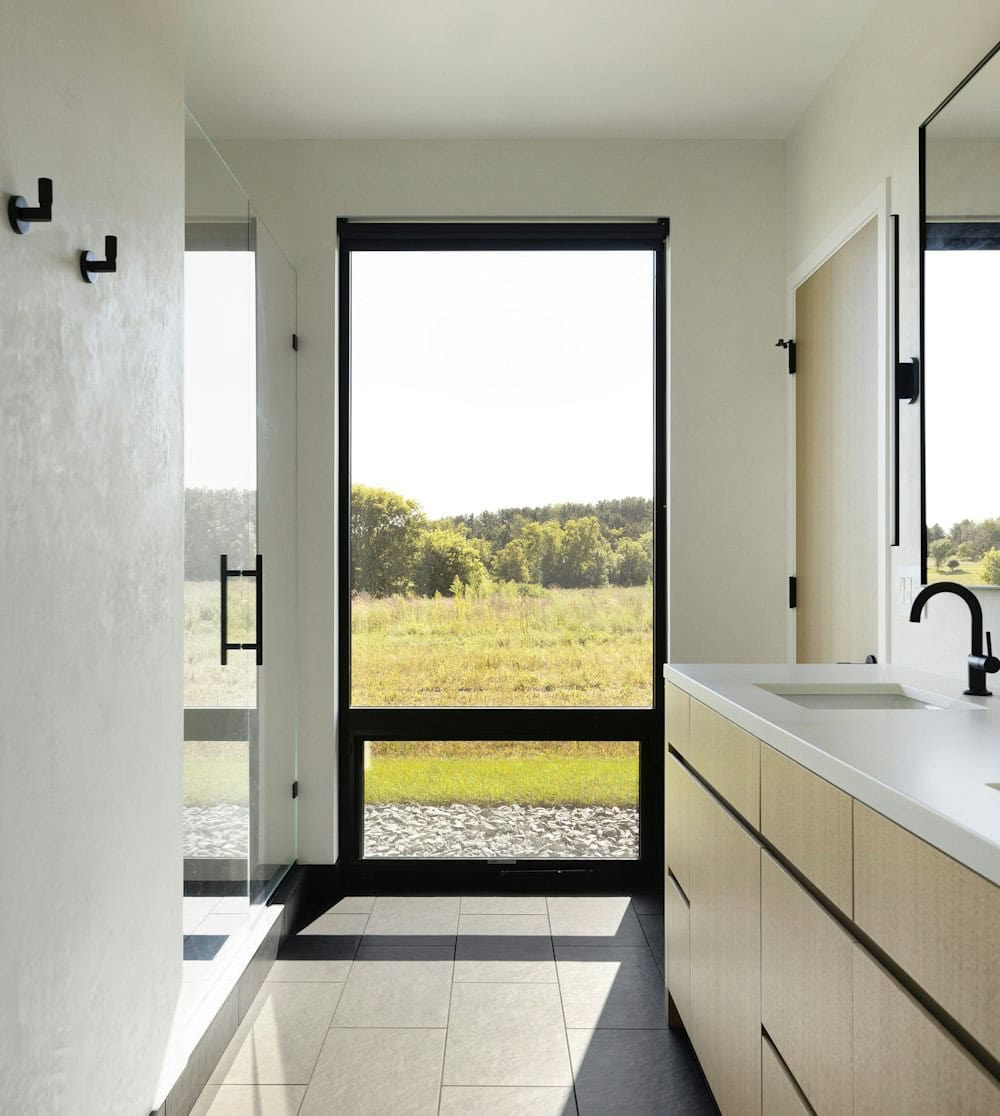 Bathroom with a tall black-framed window framing the meadow and trees, a glass shower, white oak vanity and black fixtures