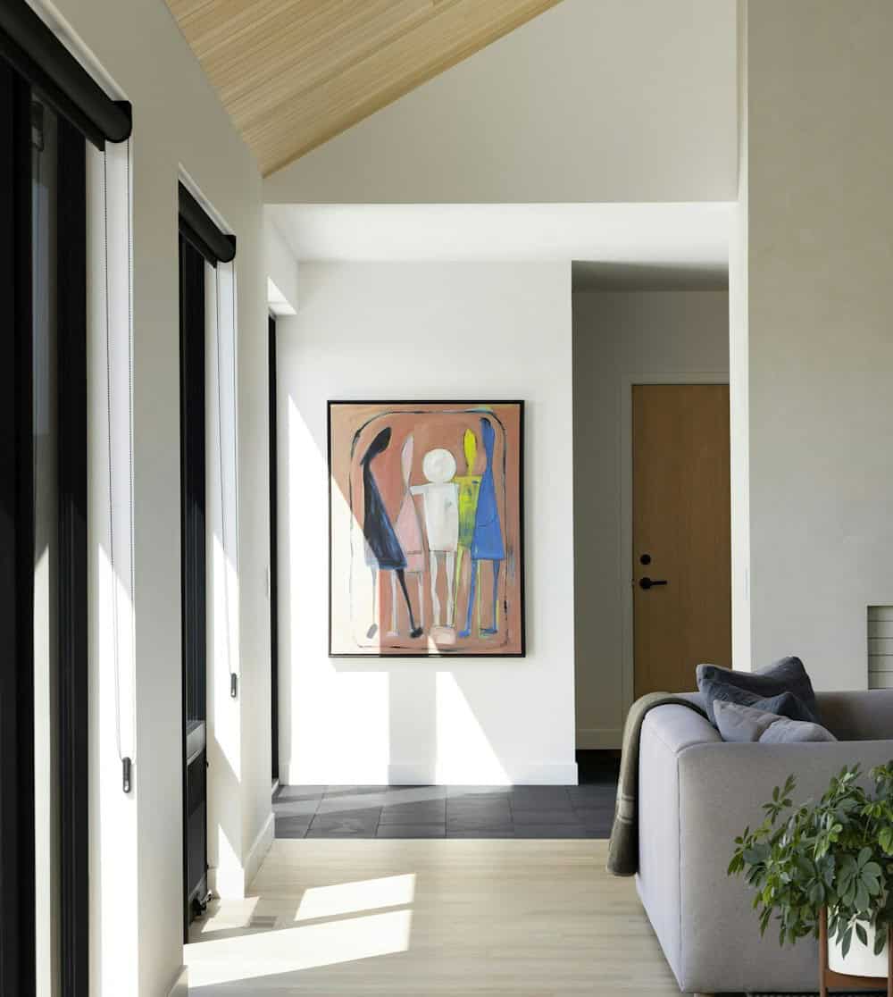Figural painting on a white wall at the end of a sunlit hallway with slate tile, a white oak door and a gray sofa in the foreground