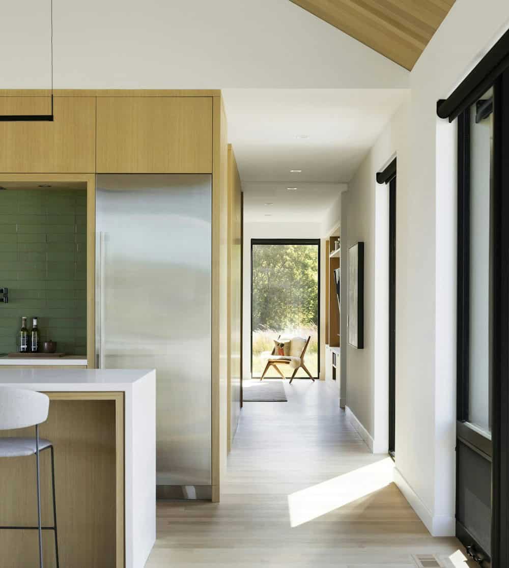 Stainless refrigerator set into white oak cabinetry beside green tile, with a light oak hallway leading to a den and a tall window