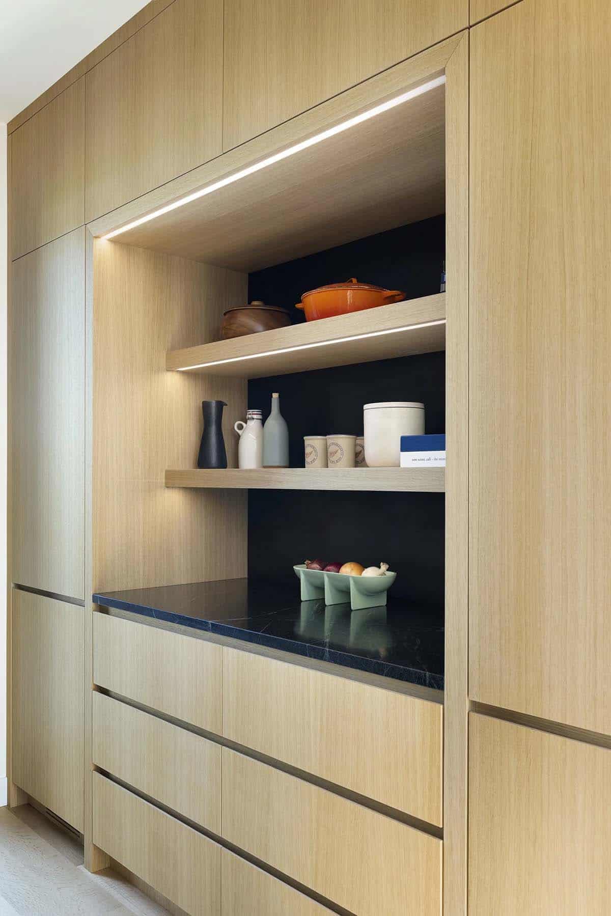 White oak built-in with lit open shelves holding pottery and an orange Dutch oven above a black stone counter and flat-front drawers