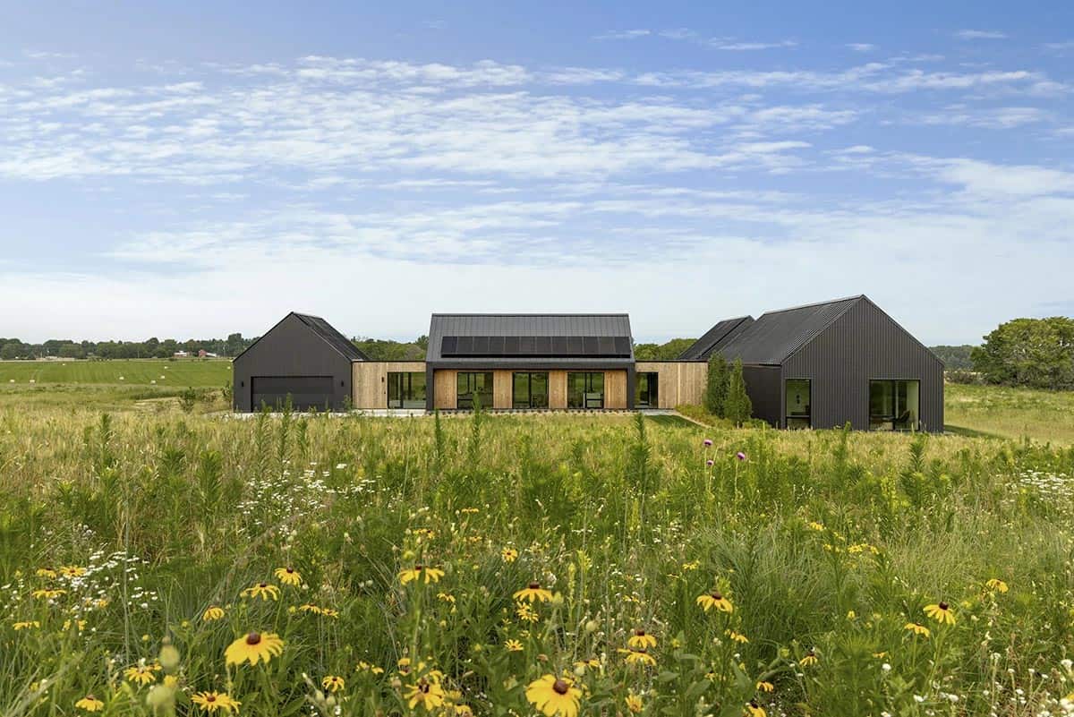 Black gabled pavilions at dusk behind a green summer meadow, with a lit covered patio, a louvered pergola and black planter boxes
