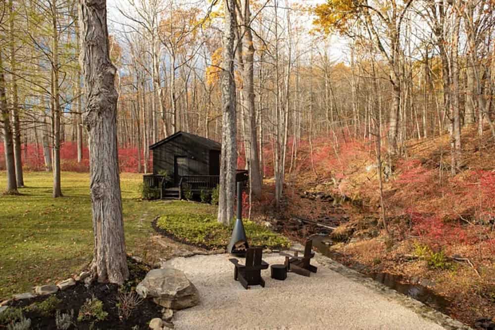 Gravel fire pit area with two Adirondack chairs and a small black cabin beside a creek