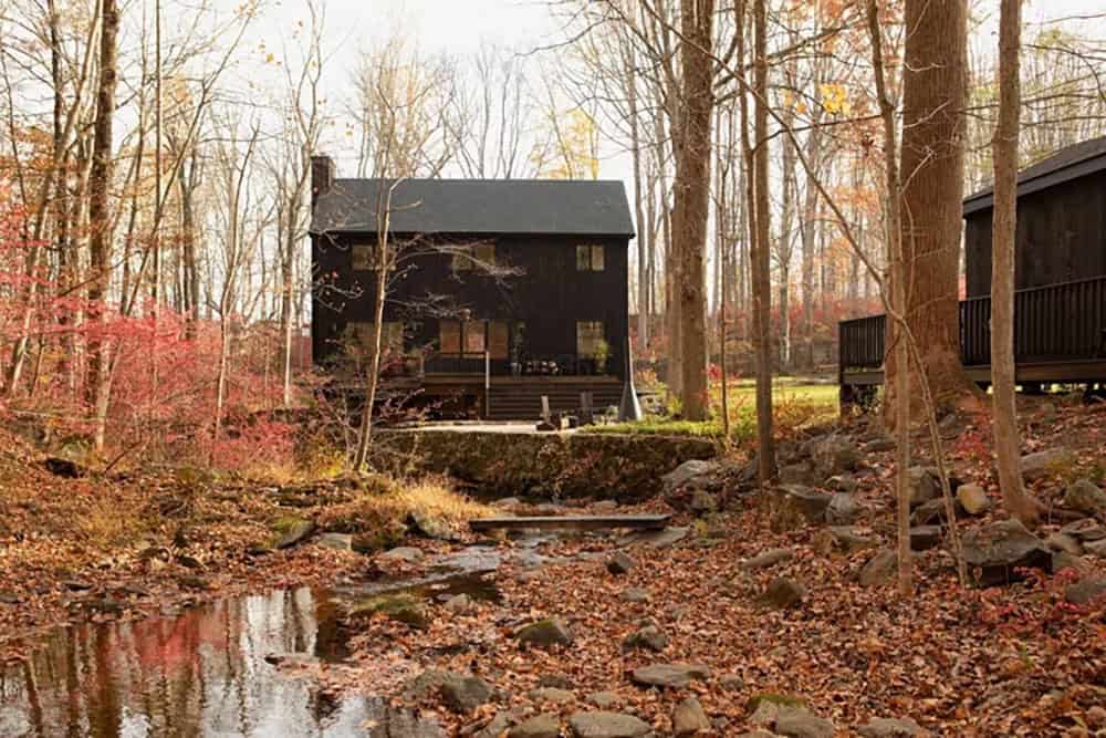 Black timber house and cabin viewed across a rocky creek bed strewn with fallen leaves