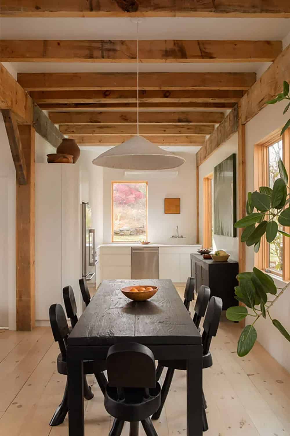 Narrow dining area with a black table, rustic ceiling beams, and a conical pendant lamp leading to the kitchen