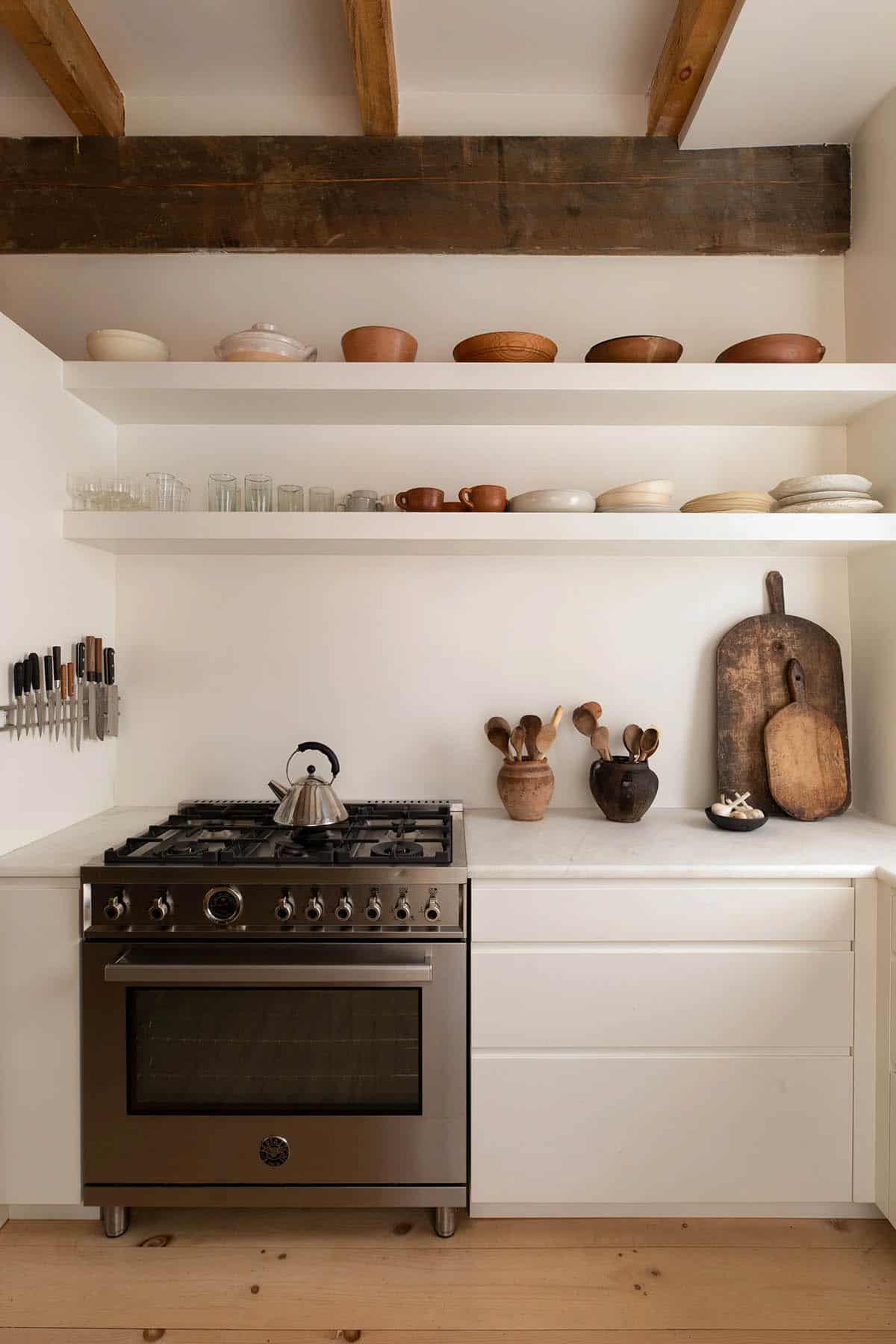 Kitchen range wall with floating white shelves holding ceramics, glassware, and wooden cutting boards