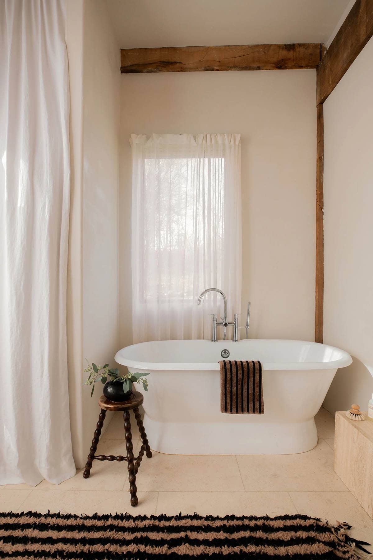 Freestanding white tub by a sheer-curtained window with a striped towel and a beaded wooden stool