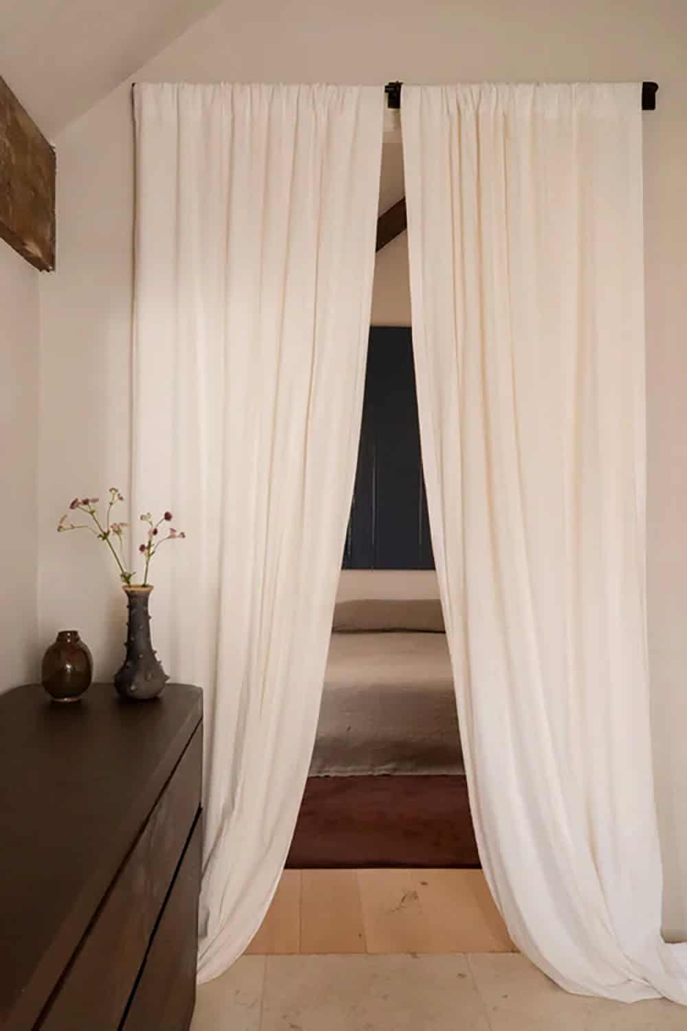 Linen curtains parted to reveal a bed and dark blue artwork beyond