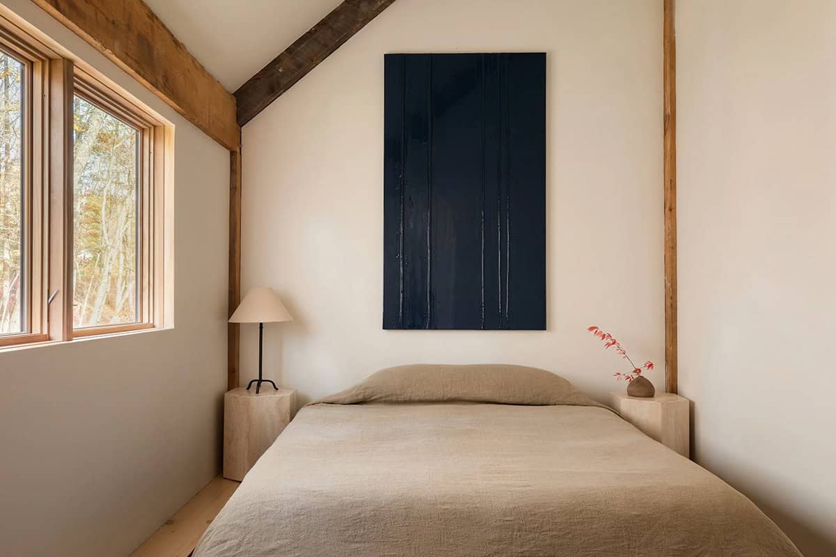 Minimal bedroom with a linen-covered bed, travertine nightstands, and a dark blue painting under a vaulted ceiling