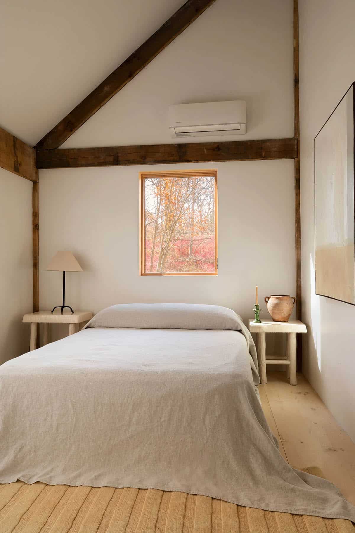Vaulted bedroom with a linen-covered bed, log-style side tables, and a small window onto autumn foliage