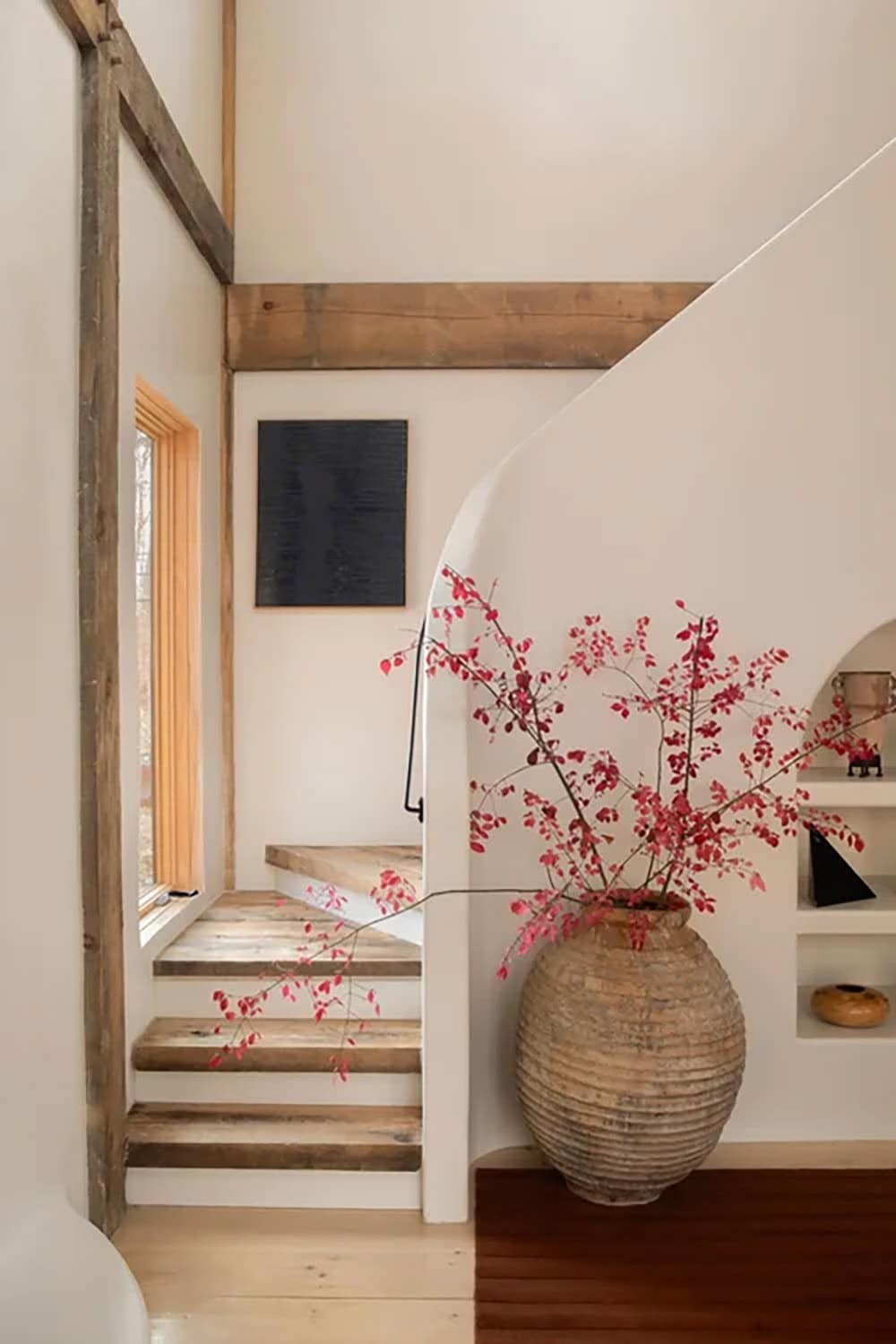 Wood-tread staircase beside a tall window, with a large urn of red branches against a curved plaster wall