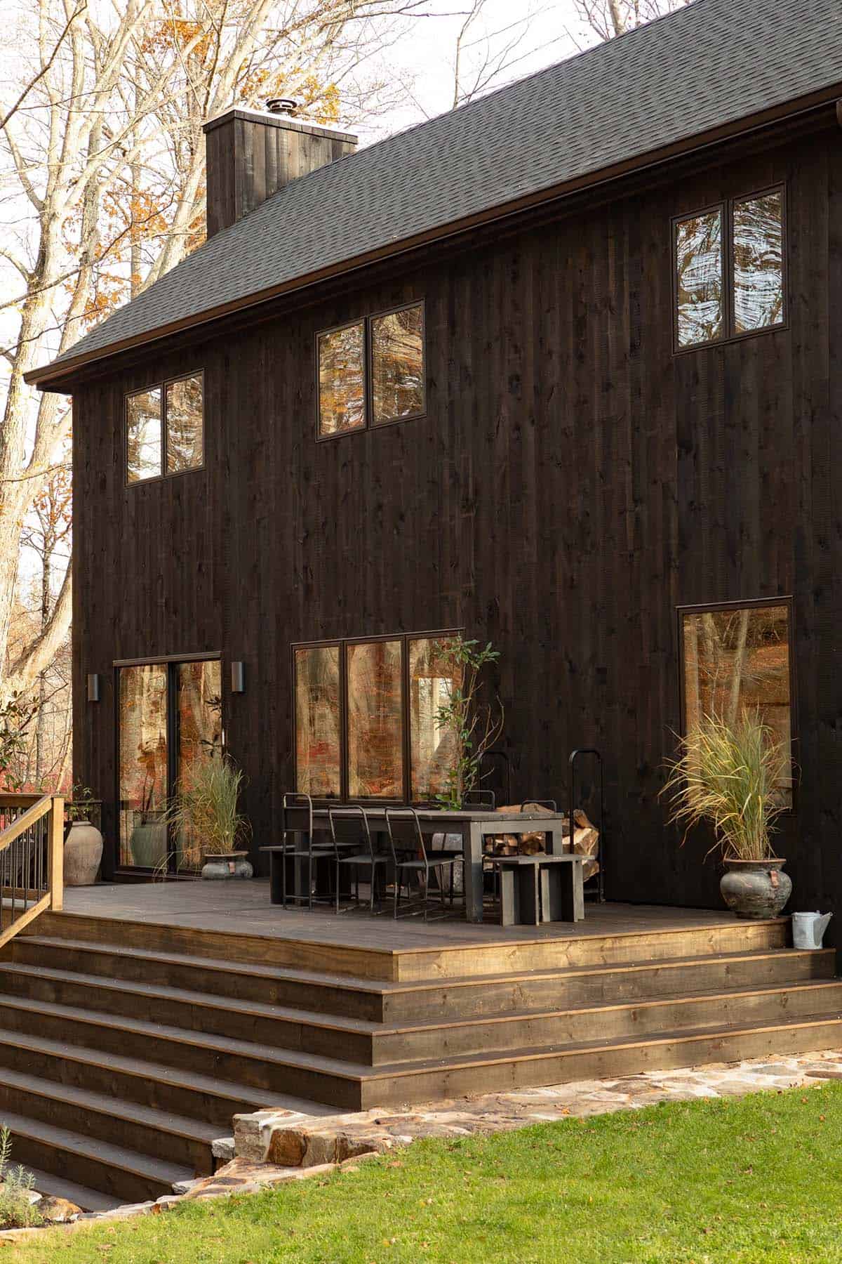 Rear of the black timber house with a deck, outdoor dining table, stacked firewood, and wide wooden steps