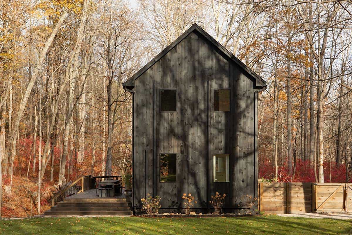 Gable end of the dark timber house with a side deck, framed by autumn woods and a wood fence