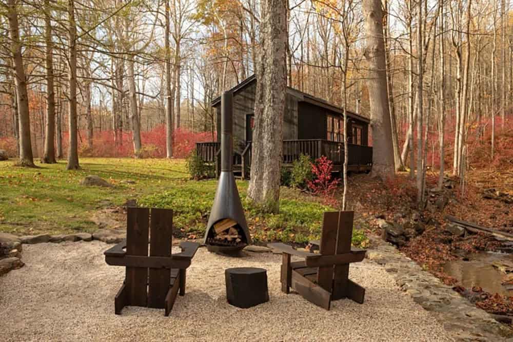 Fire pit with two dark Adirondack chairs and a log stool, with the cabin on a deck behind
