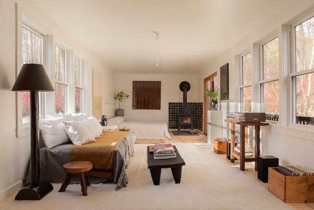 Long studio guest room with a wood stove, bed, record collection, and rows of windows onto the trees