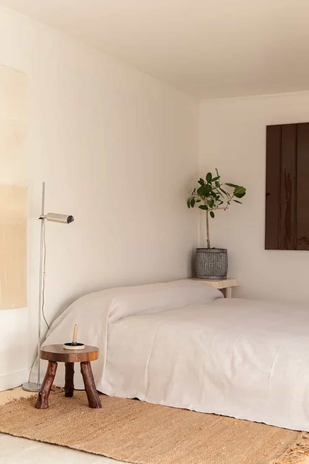 Studio bed with white linen, a wooden stool, a floor lamp, and a potted plant in the corner