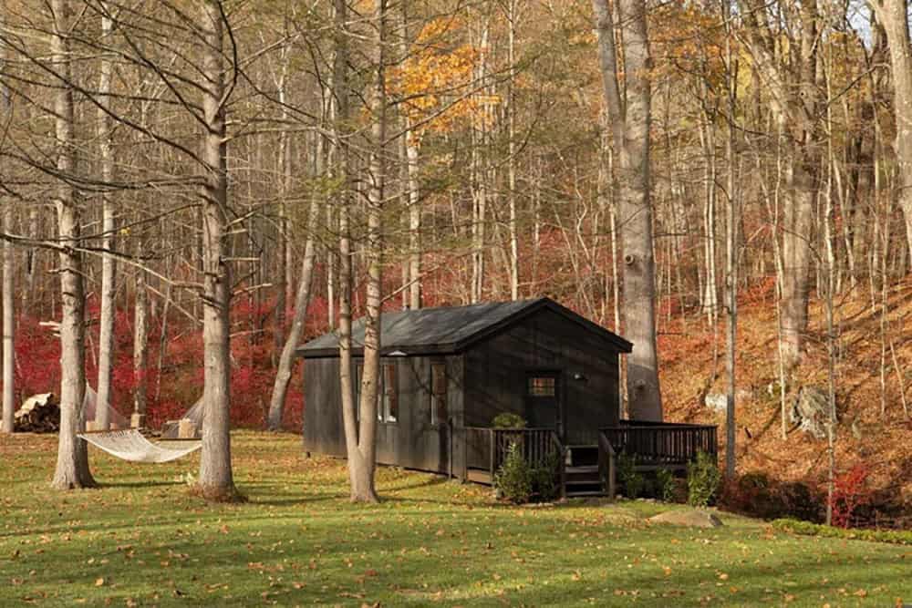 Black cabin on a lawn with a hammock between trees and a hillside of red foliage behind