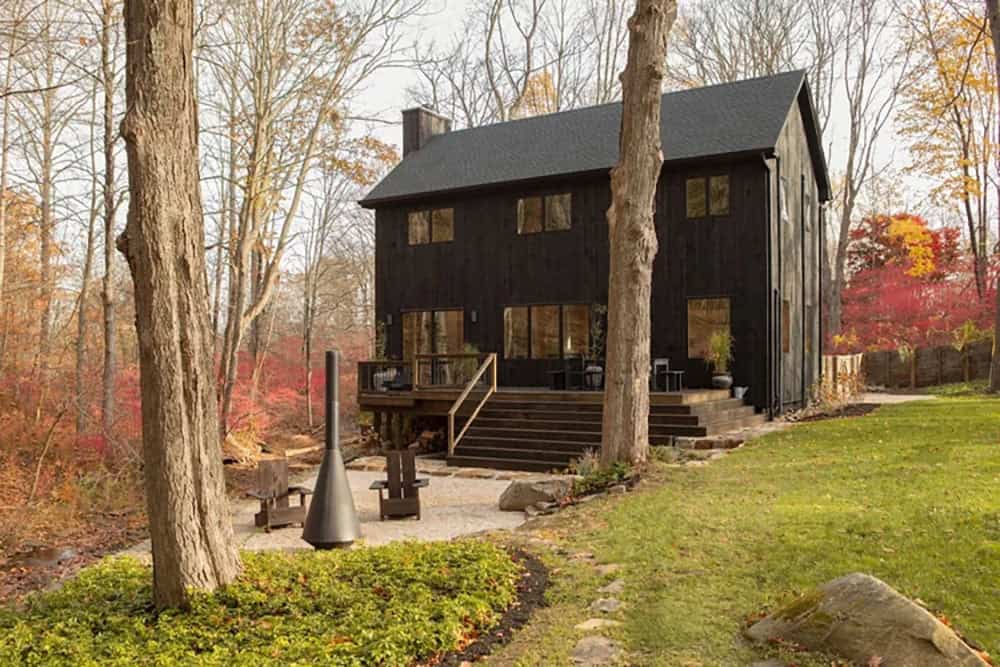 Black house beyond a gravel fire pit and lawn, with a chimney fire and Adirondack chairs