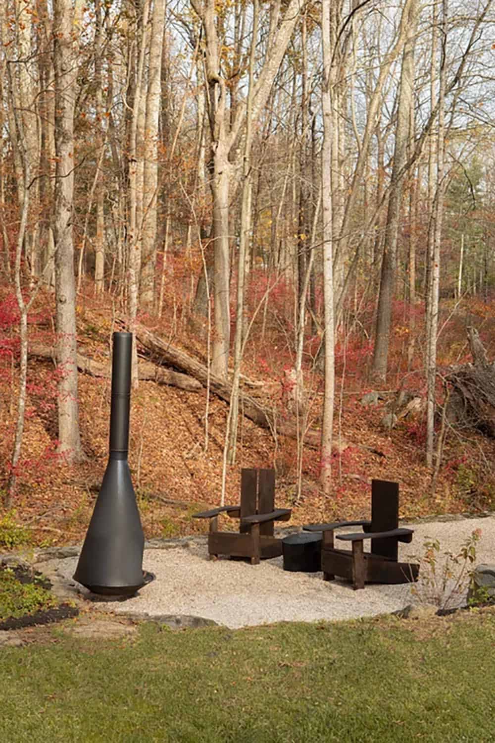 Tall black fireplace and two dark chairs on a gravel patio against a leafy hillside