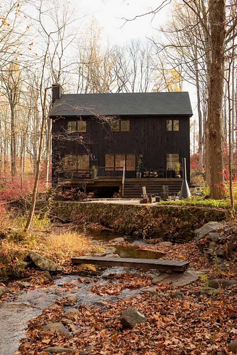 House seen across a creek, with a small plank footbridge, a stone retaining wall, and fallen leaves