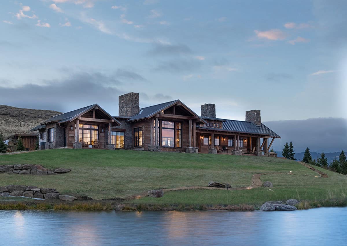 Ranch home with covered porches and stone chimneys on a grassy hillside above a pond at dusk