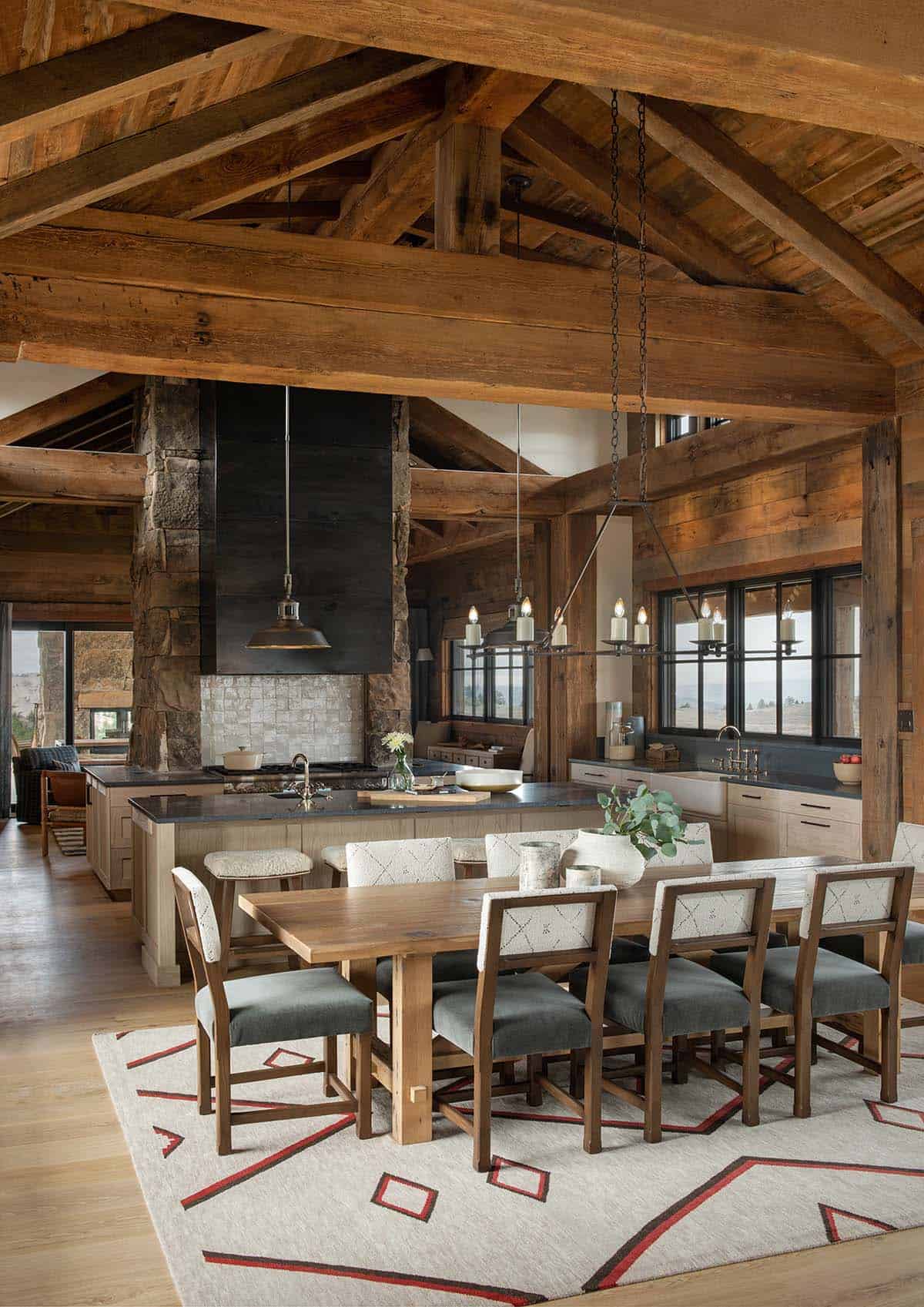 Open dining room and kitchen under reclaimed timber trusses with a long wood table, linear candle chandelier and geometric rug