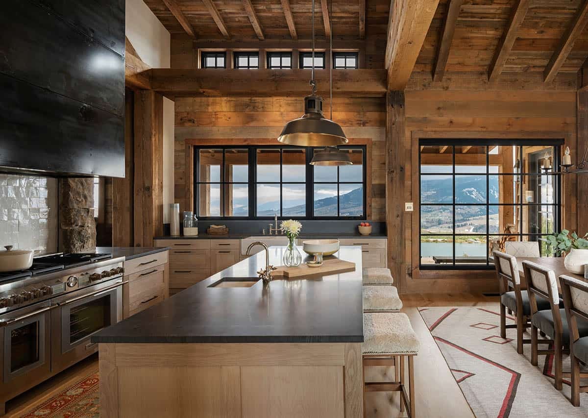 Kitchen island with a dark stone countertop and sheepskin stools, black steel range hood and windows overlooking mountains and a pond