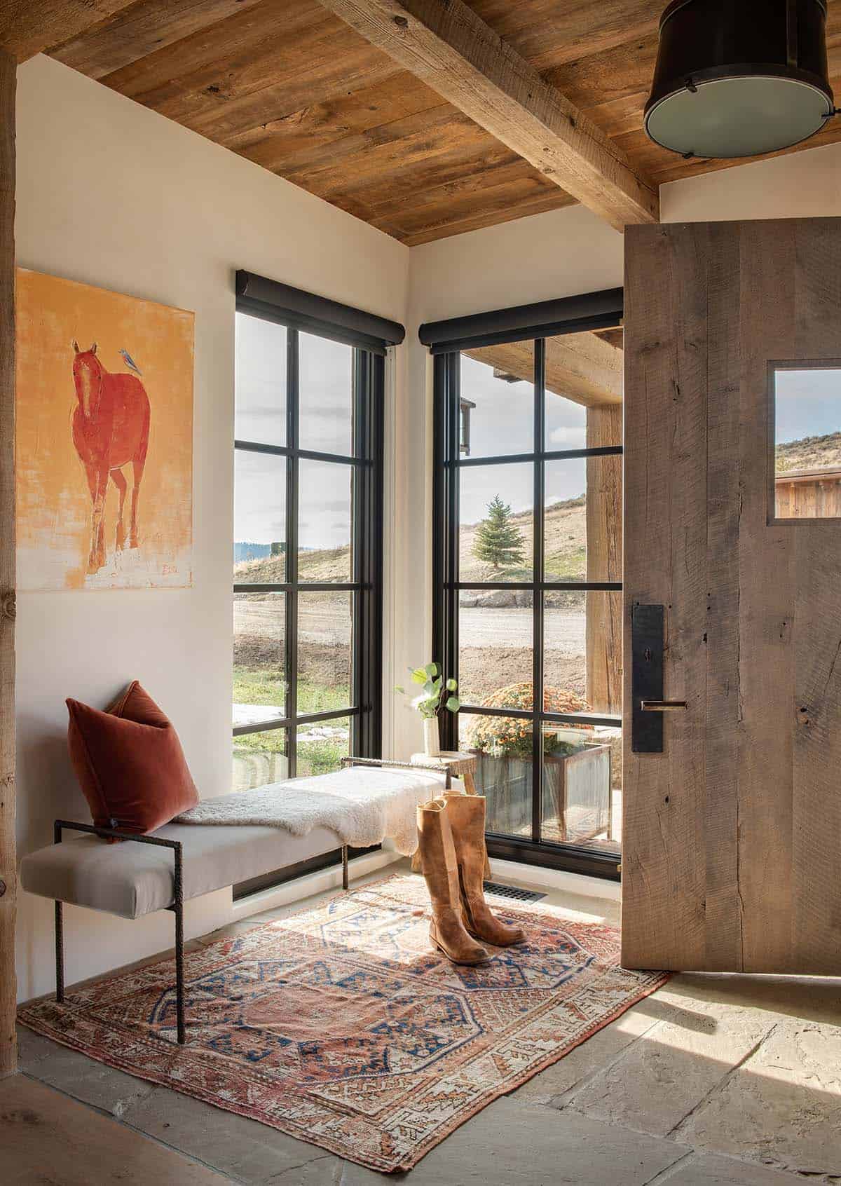 Entryway with a reclaimed wood front door, upholstered bench, red horse painting, vintage rug and black-framed corner windows