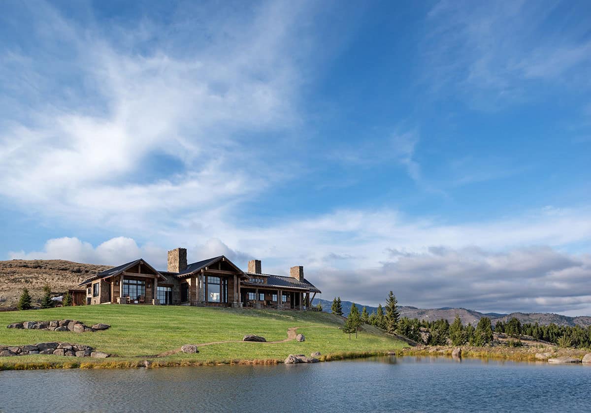 Ranch home on a green hillside above a pond, with evergreens and distant mountains under a wide blue sky