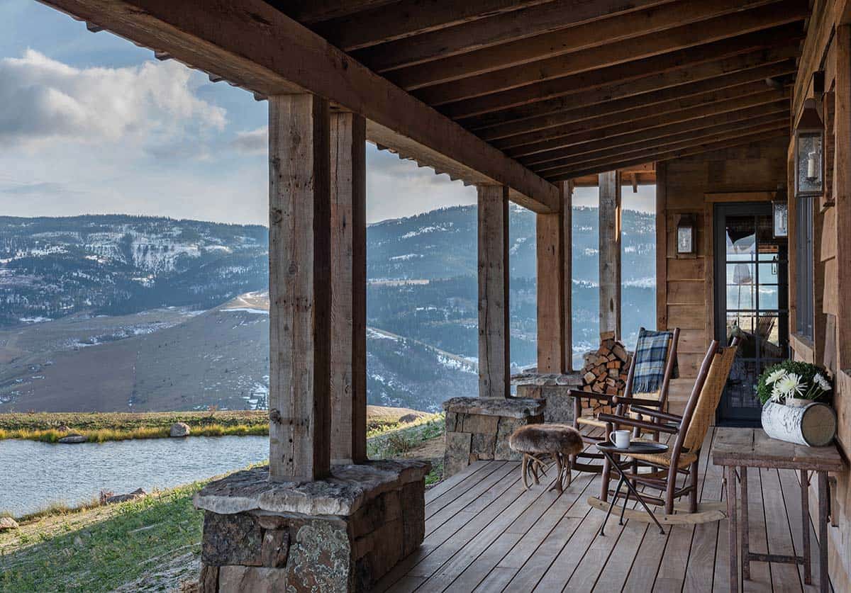 Covered porch with timber posts on stone bases, rocking chairs and a firewood stack overlooking a pond and snowy mountains
