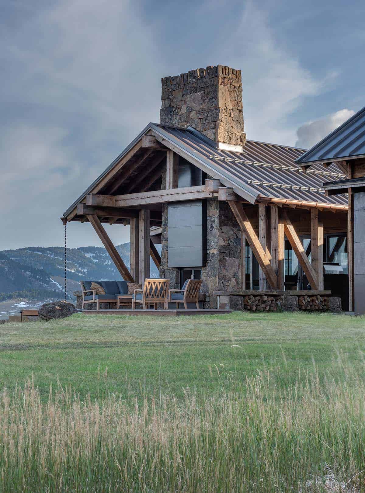 Covered outdoor living area with angled timber braces, a stone fireplace chimney and teak seating beside a grassy meadow