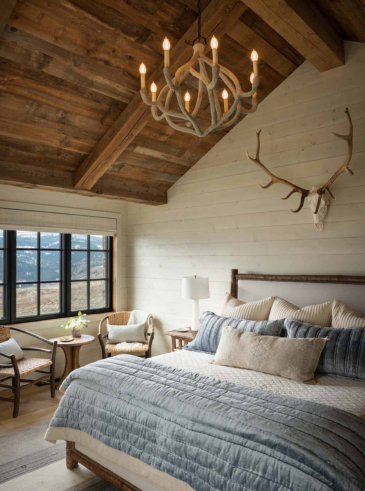 Bedroom with a wood plank vaulted ceiling, branch-style chandelier, elk antler mount, blue quilt and a window with valley views