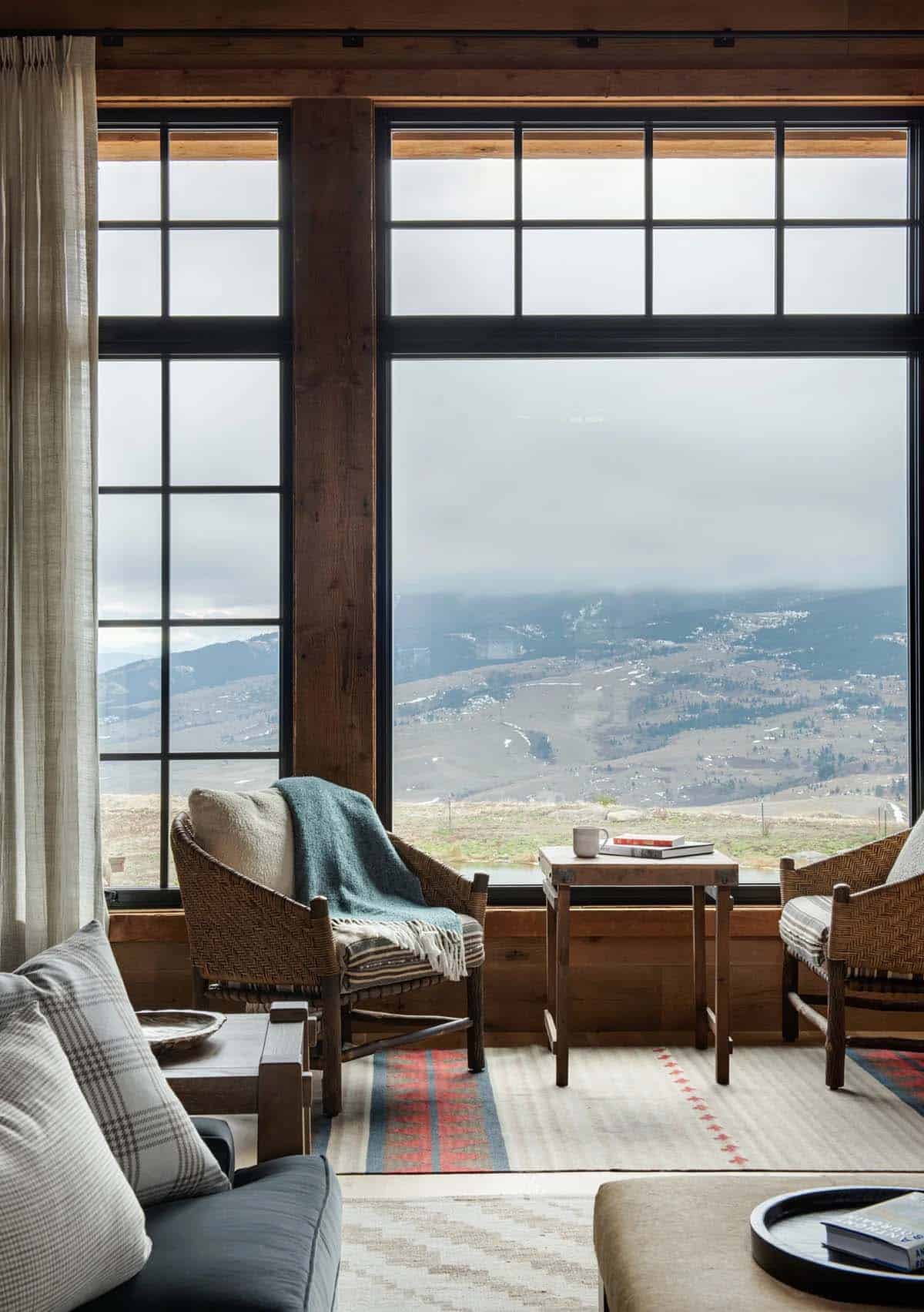 Woven armchairs and a side table in front of a large black-framed window overlooking a misty mountain valley