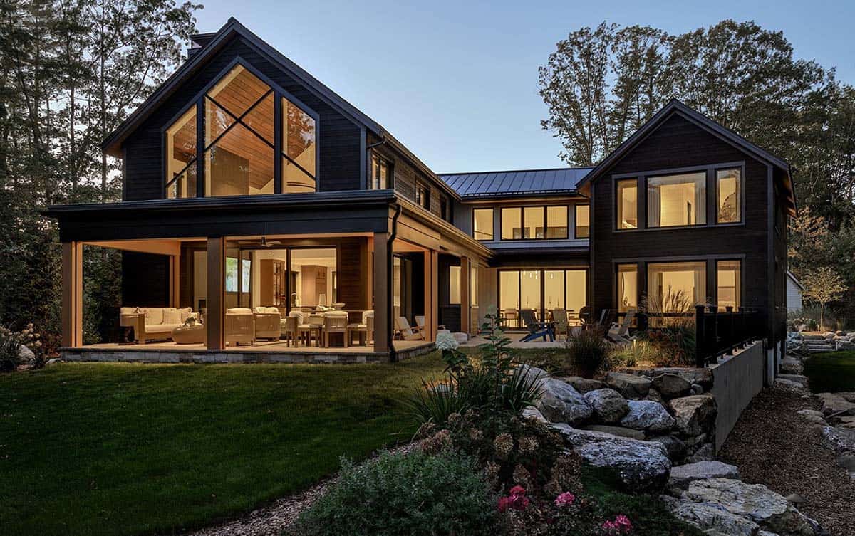 Rear of the dark charred-wood house at dusk with a glass gable window, a lit covered patio, and a boulder garden