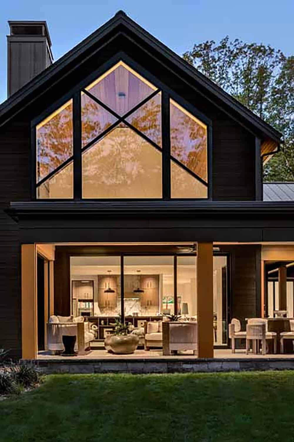 Glass gable window with crossed black mullions above a covered porch with the lit living room inside