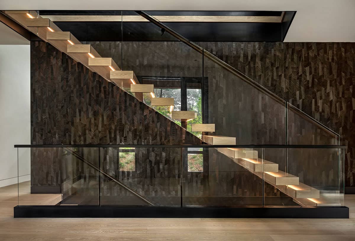 Floating oak staircase with lit treads and glass railings against a dark wood mosaic wall