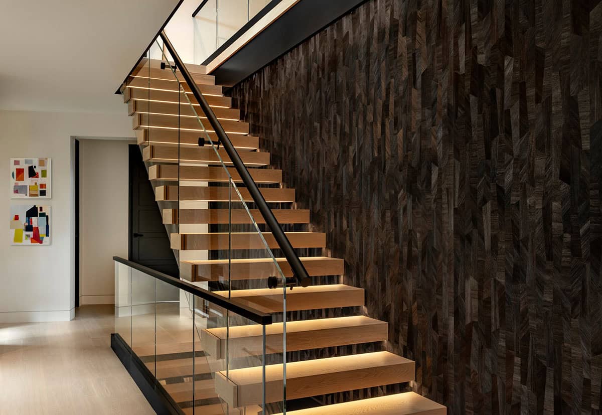 Floating oak stair treads with underlighting, a black handrail on glass panels, and a dark wood mosaic wall