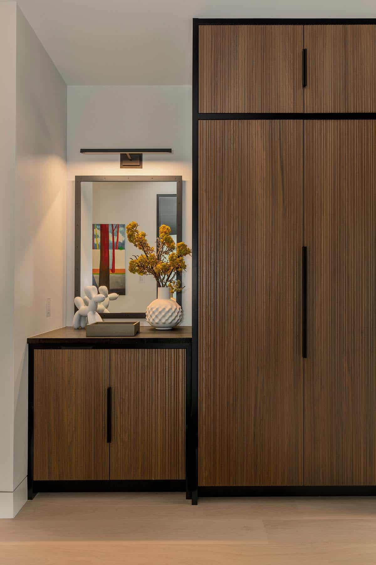 Fluted walnut cabinets with black frames beside a small stone counter with a mirror and picture light