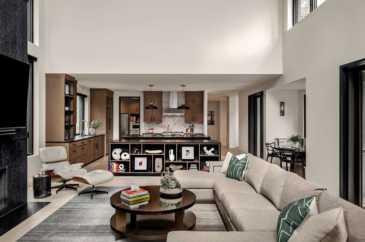 Double-height living room with a sectional and Eames lounge chair, looking past a black bookcase to the kitchen