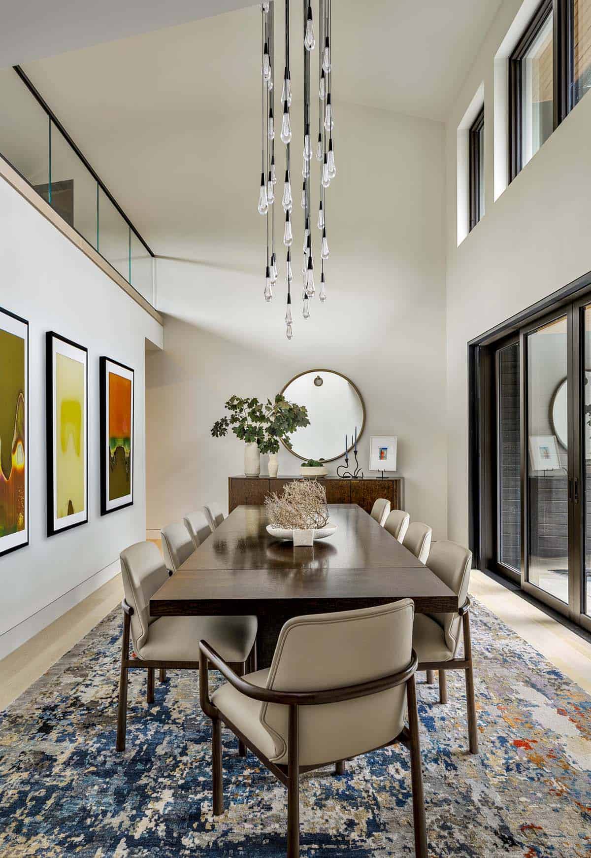 Double-height dining room with a glass drop chandelier, three abstract prints, and a blue patterned rug