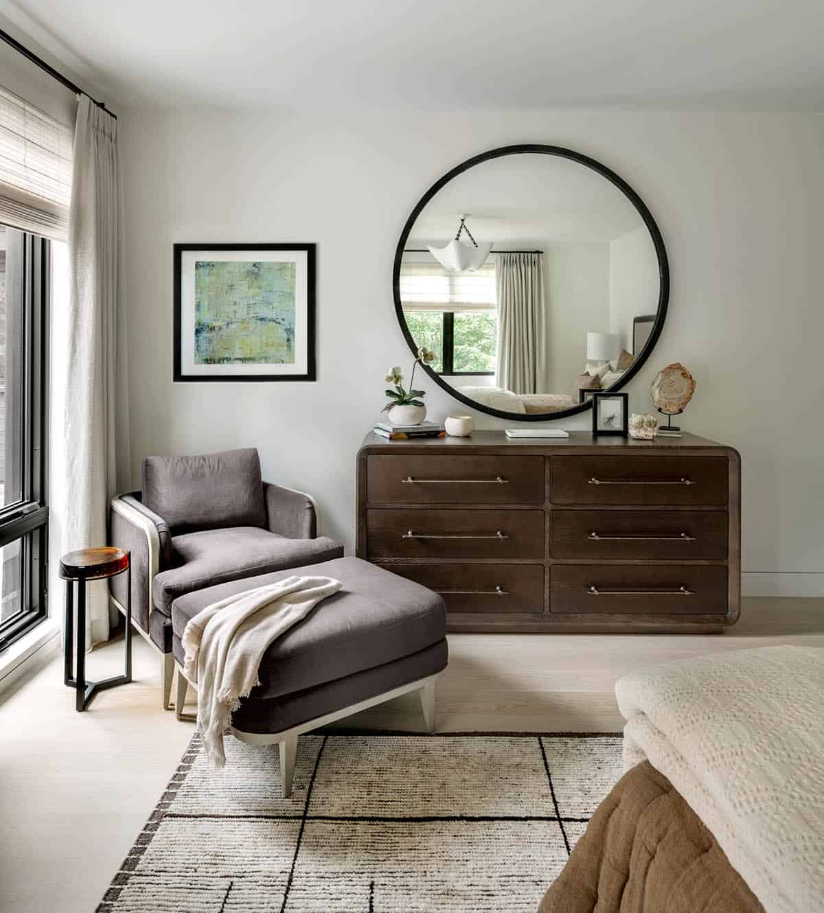 Bedroom corner with a round black mirror over a dark oak dresser, and a gray armchair and ottoman