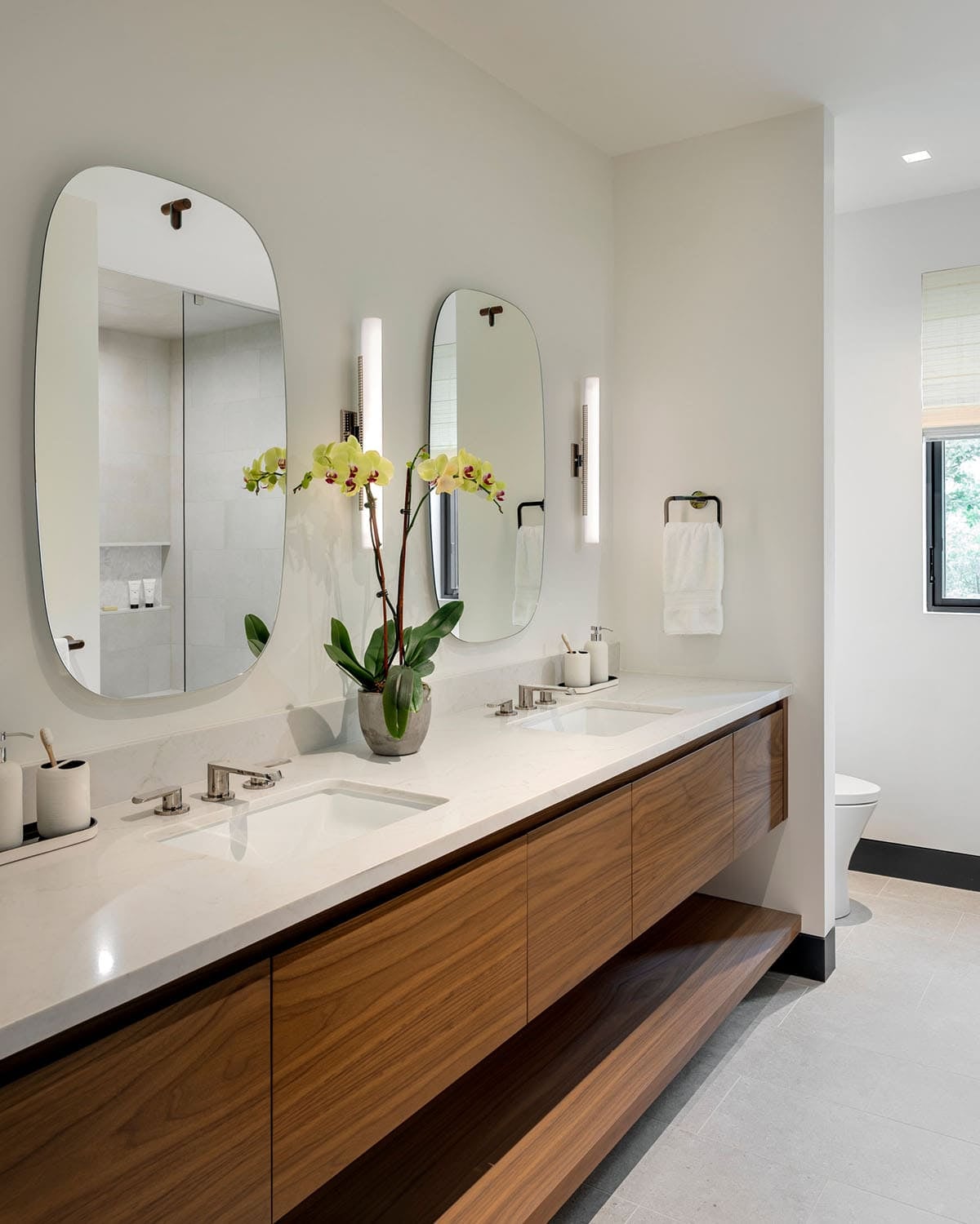 Bathroom with a floating walnut double vanity, two rounded mirrors, and a yellow orchid