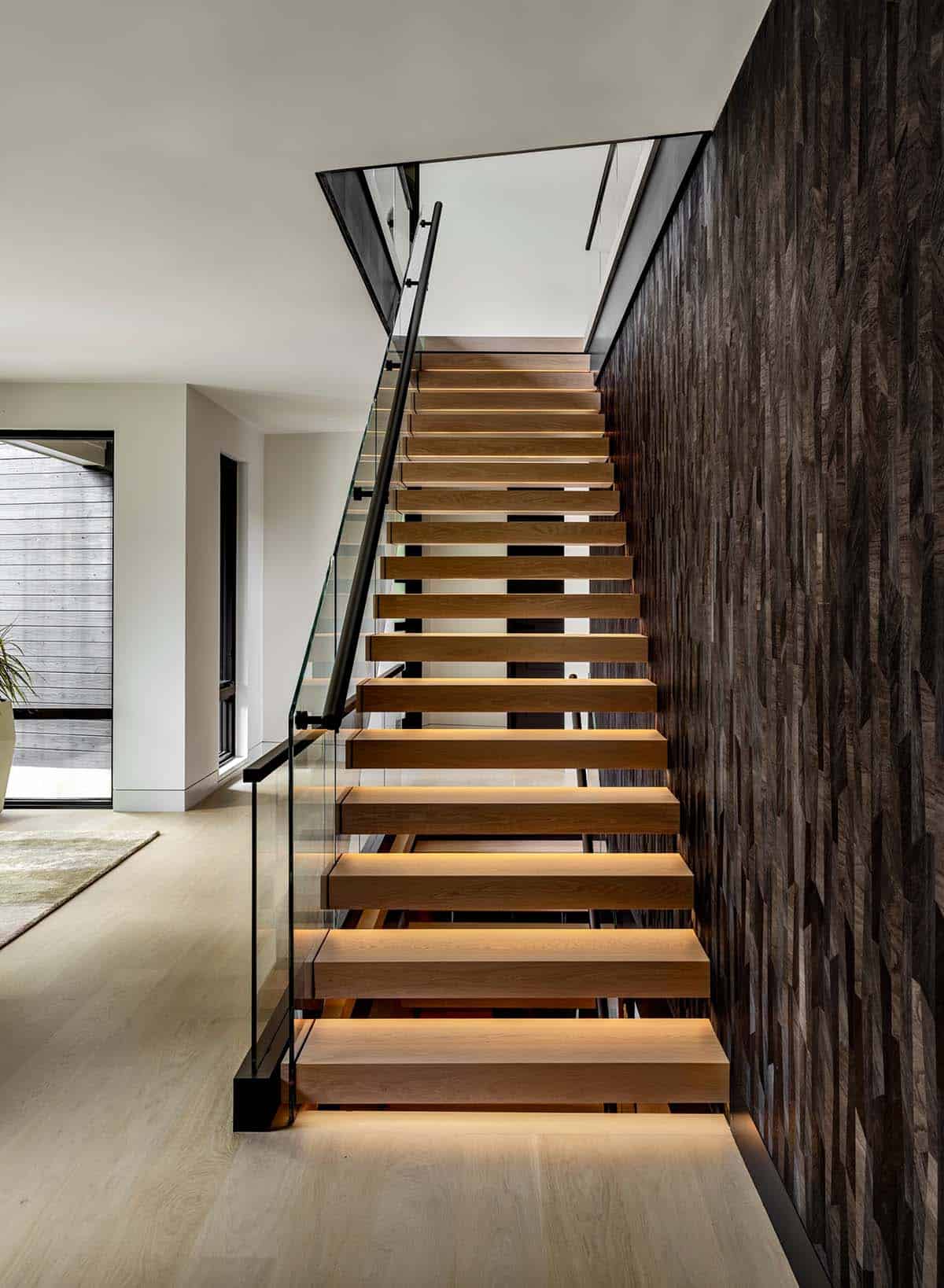 Floating oak staircase with lit treads rising beside a dark wood mosaic wall