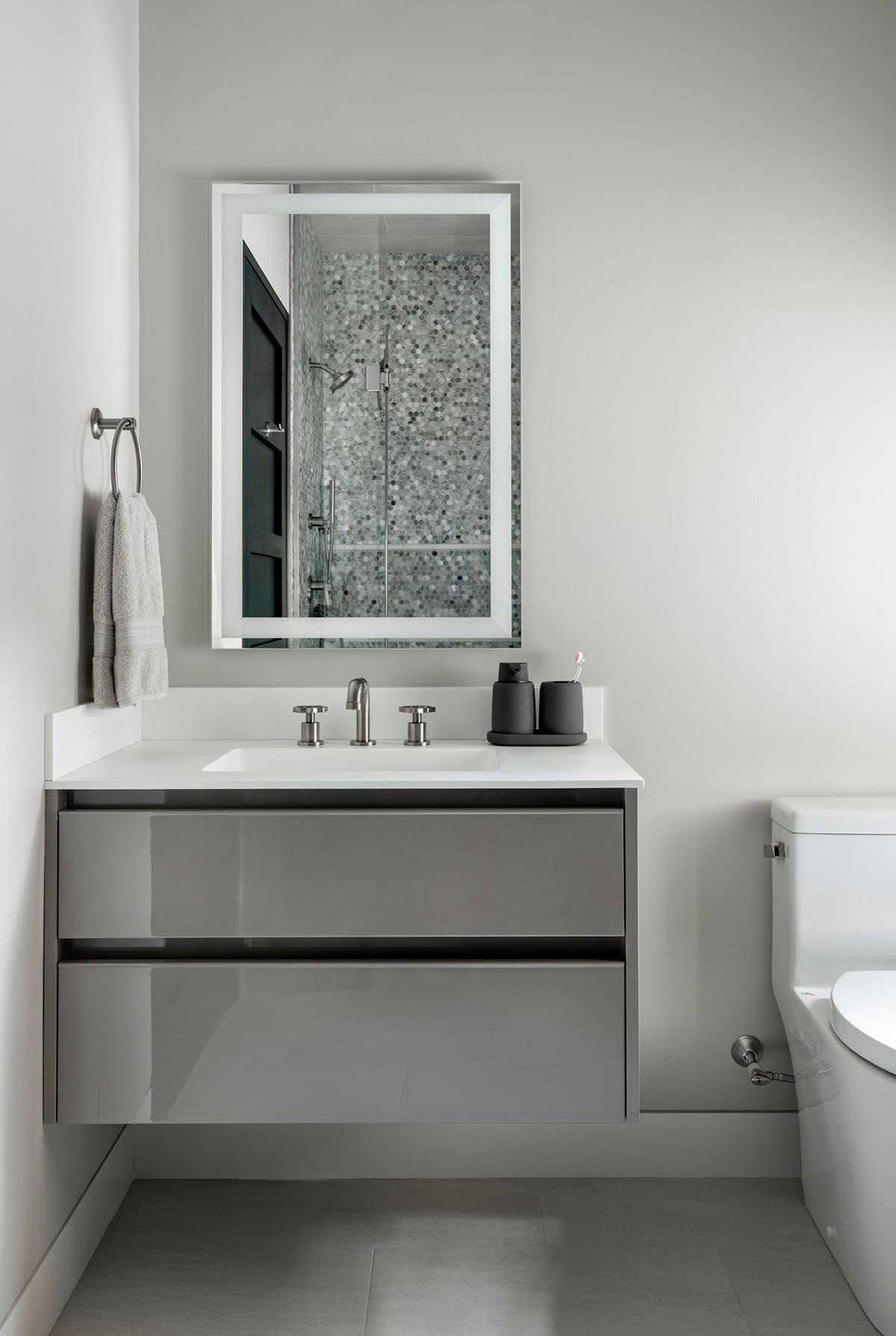 Glossy gray floating vanity under a lit mirror reflecting a mosaic tile shower