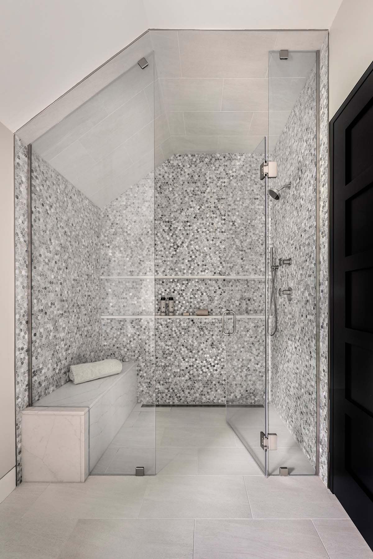 Shower under a sloped ceiling with gray hexagon mosaic walls, a stone bench, and glass doors