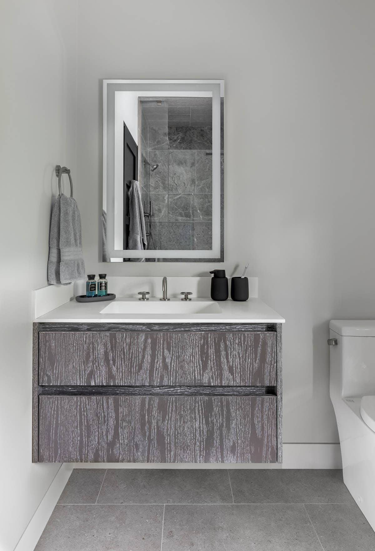 Floating cerused gray oak vanity under a lit mirror reflecting a marble tile shower