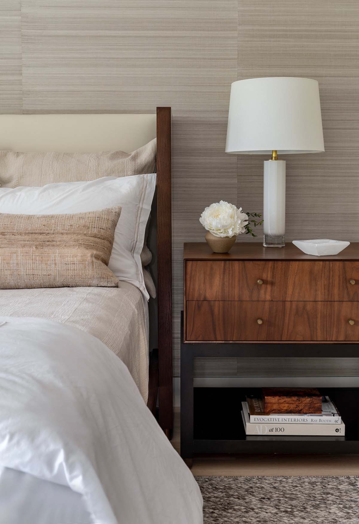 Walnut nightstand with a white lamp, a peony, and stacked books beside the bed