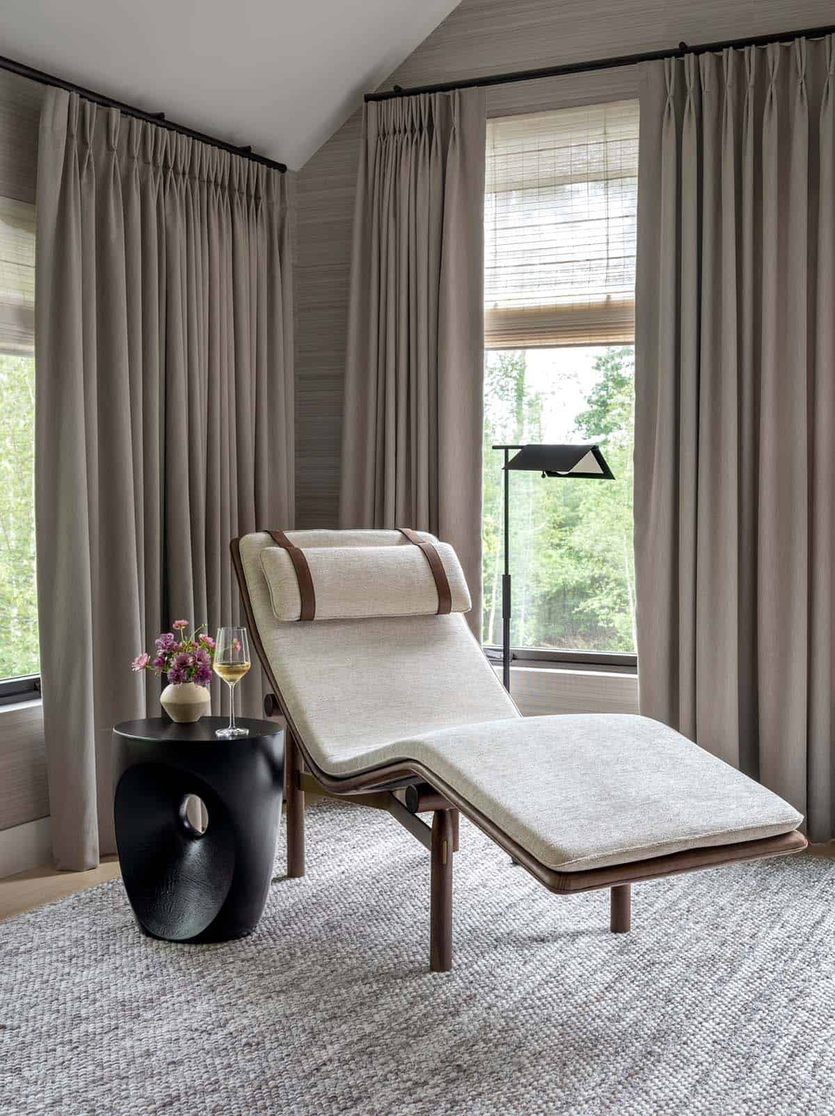 Upholstered chaise lounge with leather straps and a black side table between curtained windows
