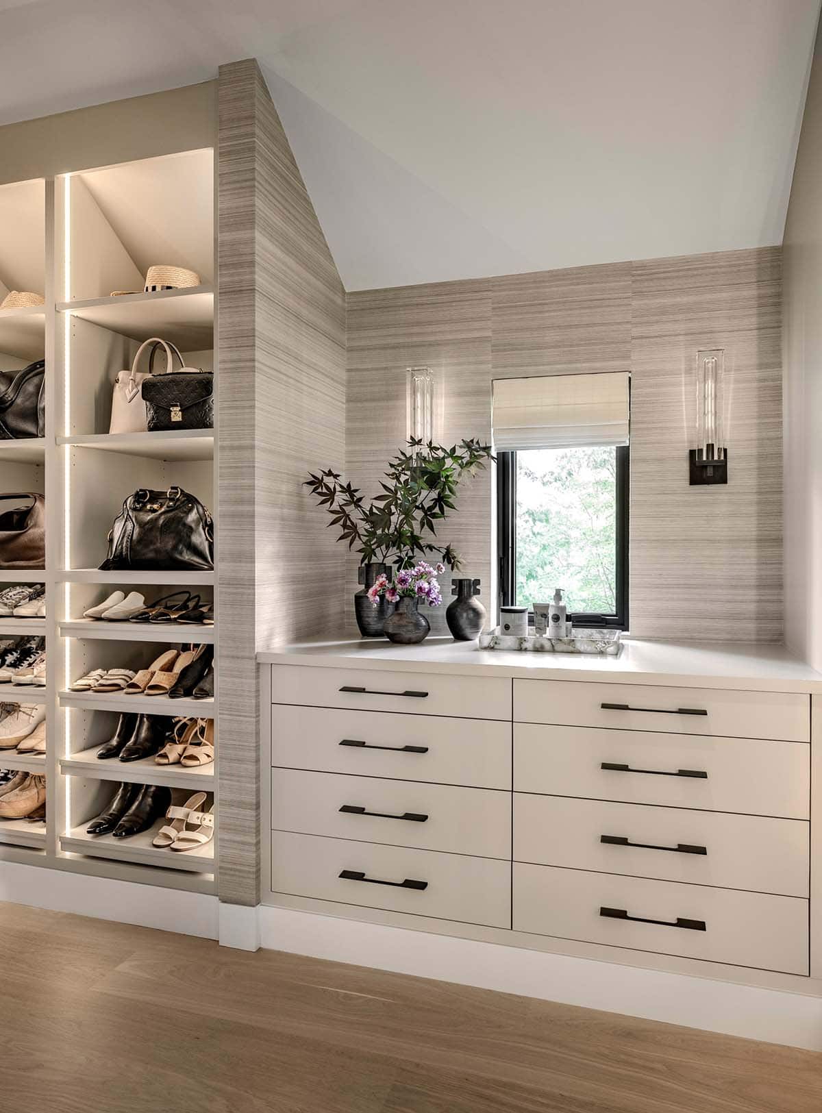 Walk-in closet with lit shelves of shoes and handbags beside white drawers under a window
